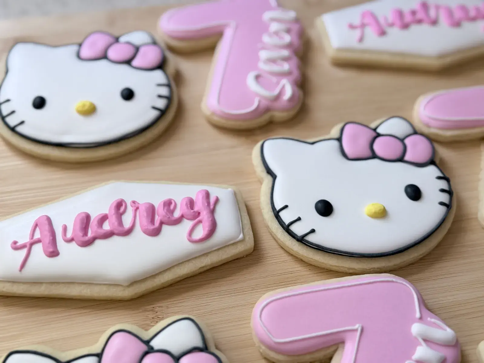 Hello Kitty birthday cookies in pink and white, including two Hello Kitty face cookies with pink bows, a hexagon plaque iced with the name Audrey in pink script, and pink numeral 7 cookies.