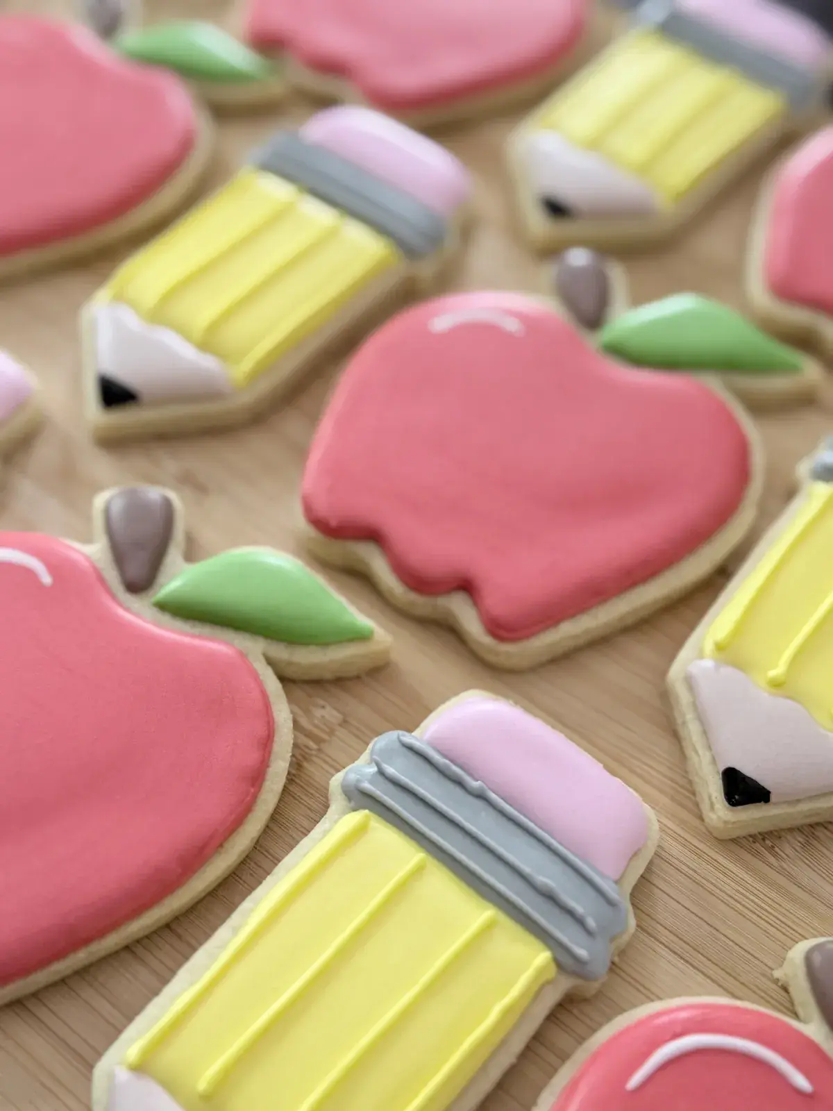 Iced sugar cookies shaped like apples, pencils, and paintbrushes in pink, yellow, and green, arranged on a wood board for a back-to-school treat set.