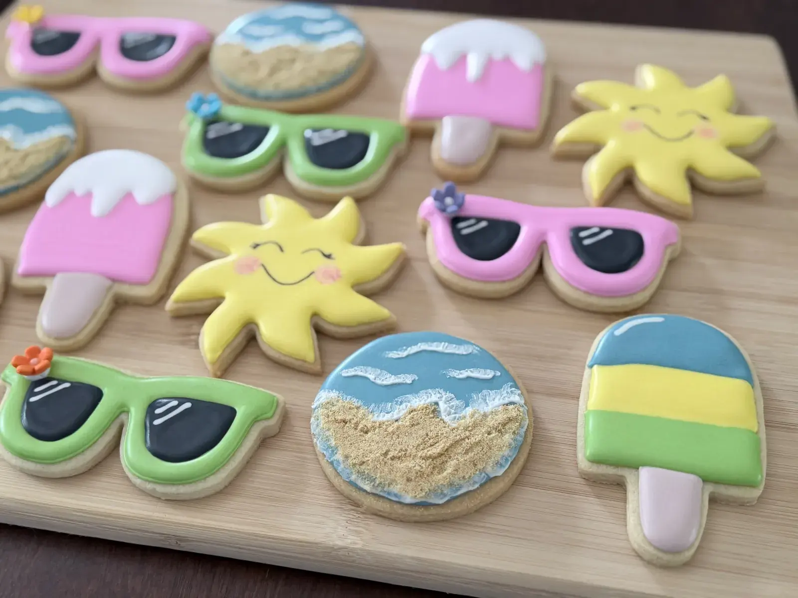 Assorted summer-themed sugar cookies: pink and green sunglasses, a smiling yellow sun, a sandy beach scene, and striped popsicles, iced in pastel colors.