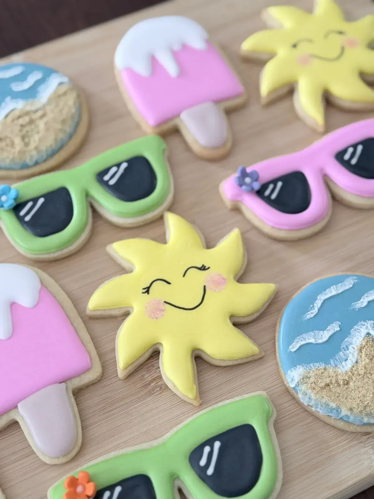 Close-up of the same summer sugar cookie set, showing a smiling sun face with rosy cheeks between green and pink sunglasses cookies and a beach-scene cookie with white wave detailing.