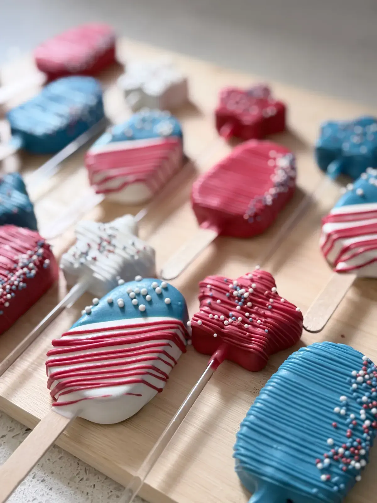 Red, white, and blue chocolate-covered cakesicles and star-shaped pops on wooden sticks, decorated with piped stripes and nonpareil sprinkles for a patriotic dessert table.