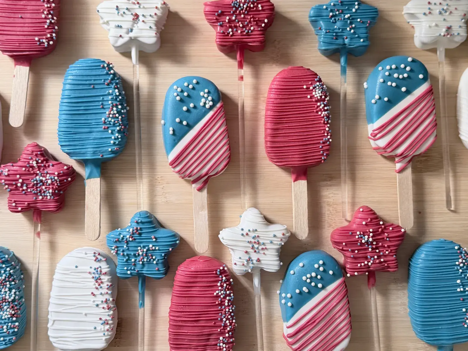 An overhead view of the same patriotic dessert spread, showing rows of red, white, and blue cakesicles and stars each finished with drizzled icing and sprinkles.