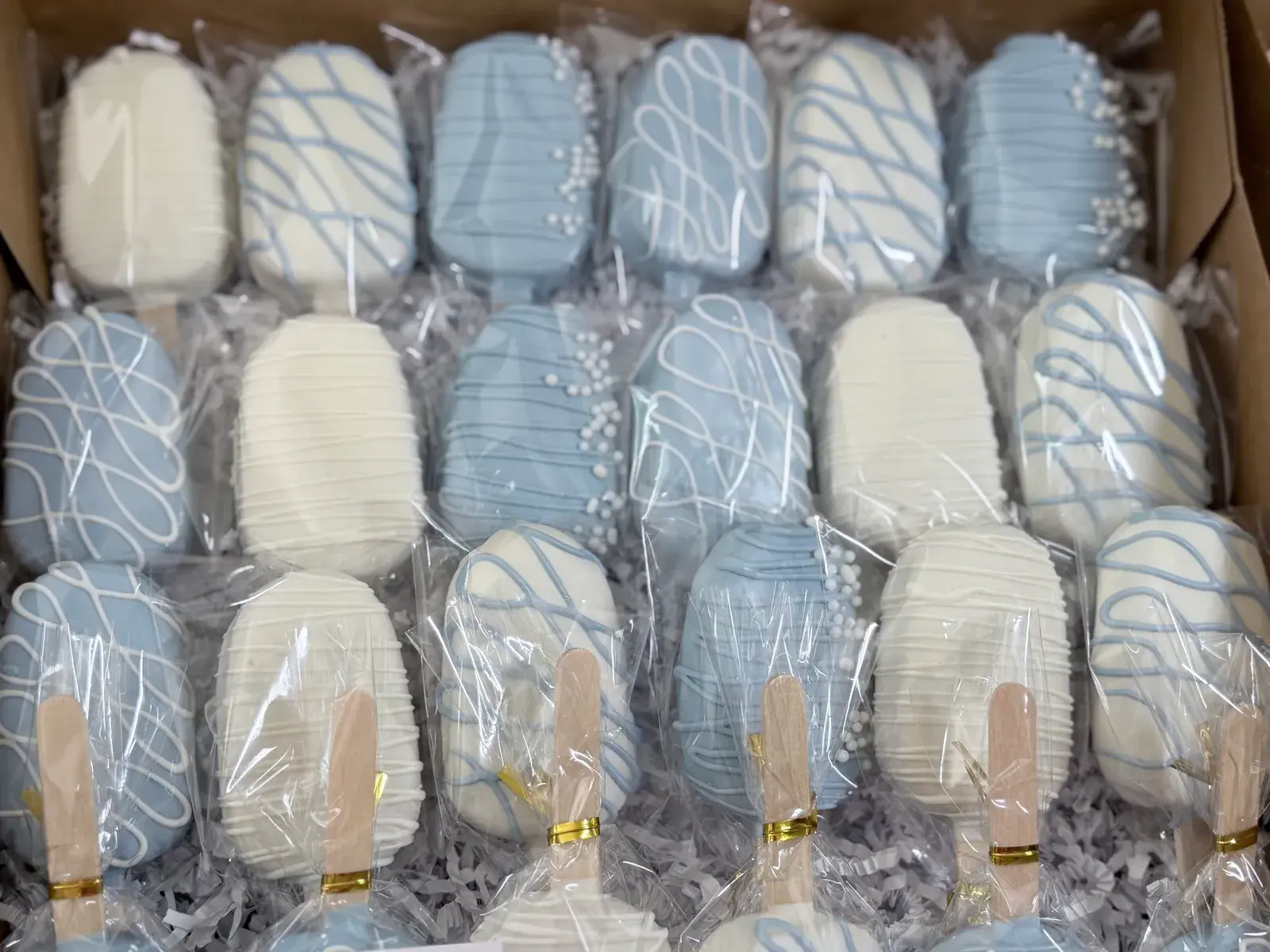 A gift box packed with white and powder-blue chocolate-dipped cakesicles, each finished with drizzled icing or a cluster of white nonpareils, wrapped individually in cellophane.