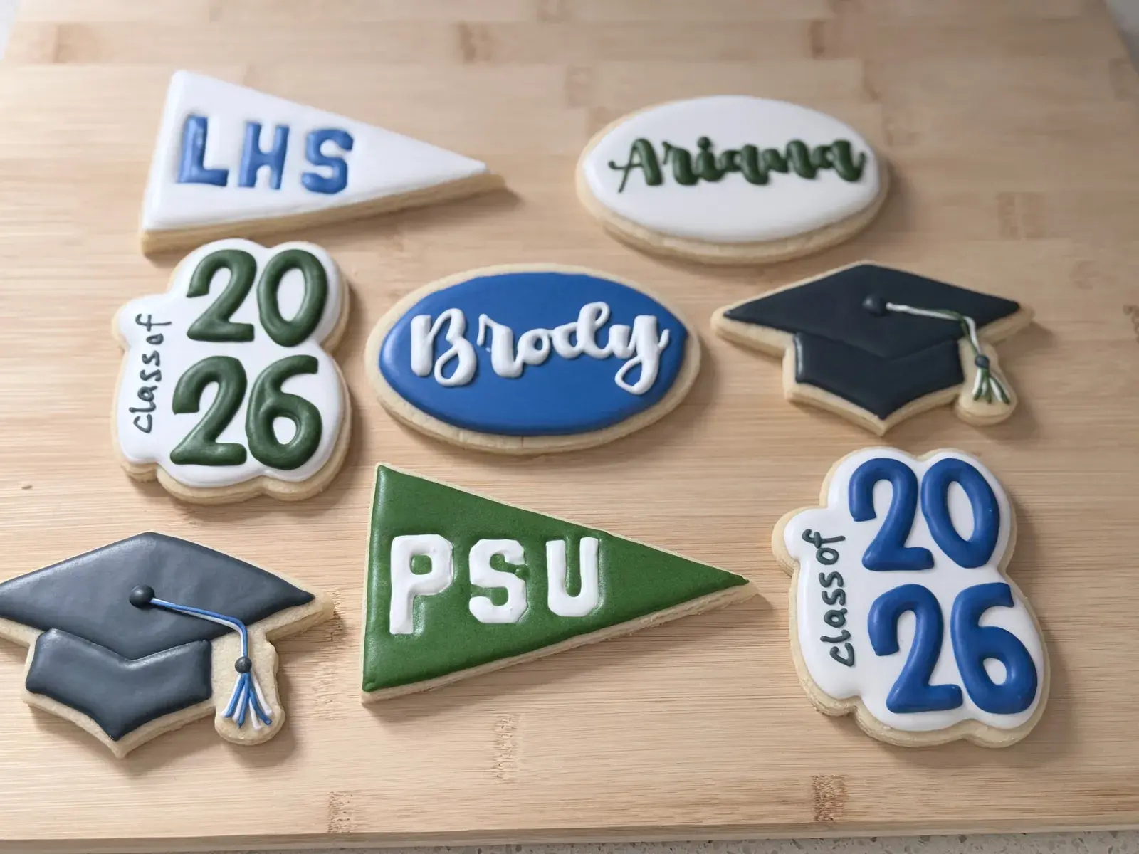 A high school graduation cookie set: a white LHS pennant, a green PSU pennant, black graduation caps with tassels, Class of 2026 plaques in green and blue, and oval cookies reading Ariana and Brody.