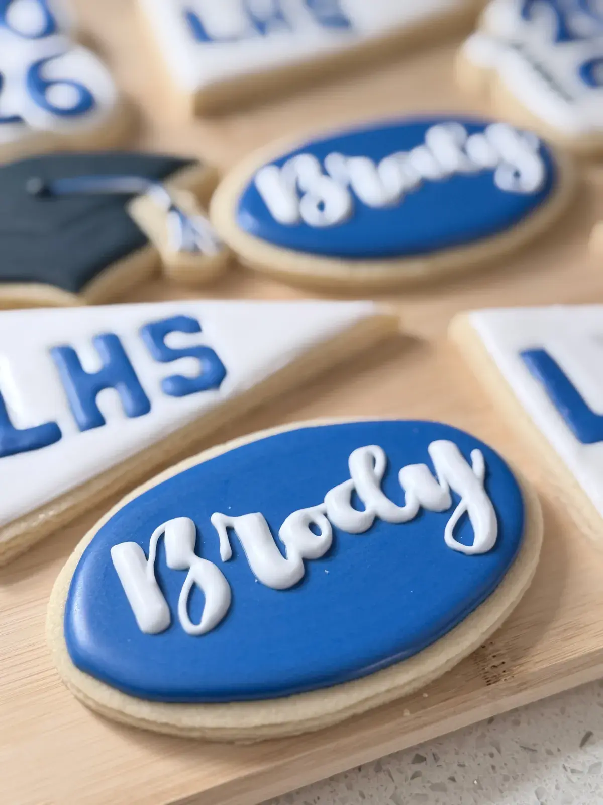A close-up of the blue oval cookie reading Brody in white script, with the LHS pennant and a graduation cap cookie blurred in the background.