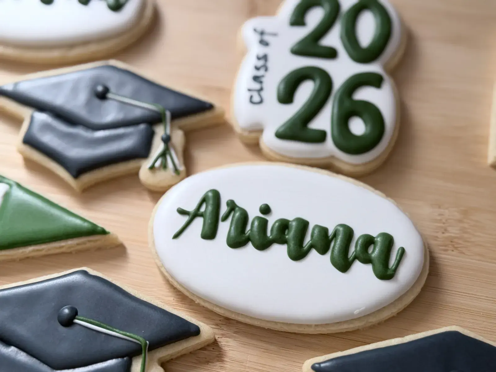 A close view of the graduation cookie set on a wood board: a large white oval iced with the name Ariana in dark green script, a white plaque reading Class of 2026 in green, charcoal graduation caps with tassels, and the corner of a green pennant.