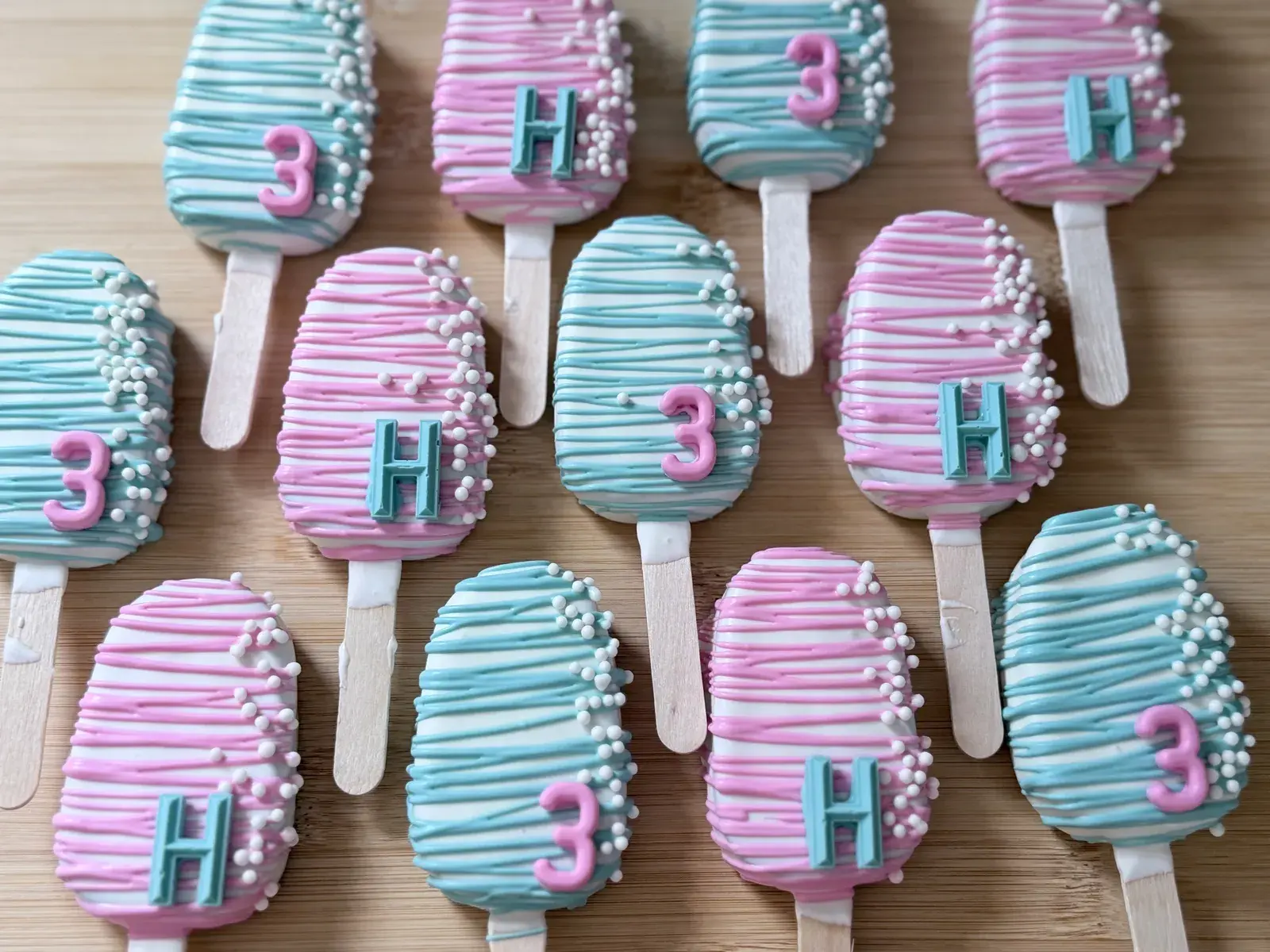 Rows of cakesicles on wooden sticks, coated in white chocolate and drizzled with pink or teal stripes, each topped with a fondant letter H or numeral 3 in the contrasting color and a scatter of white nonpareil pearls.