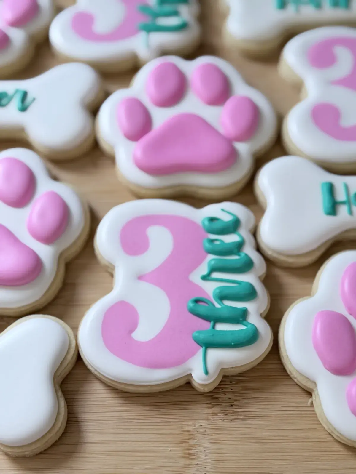 Dog-paw and dog-bone iced sugar cookies in pink, white, and teal: pink-and-white paw prints, a bone reading Harper in teal script, and a pink number 3 cookie with Three iced beside it in teal, arranged on a wood board.