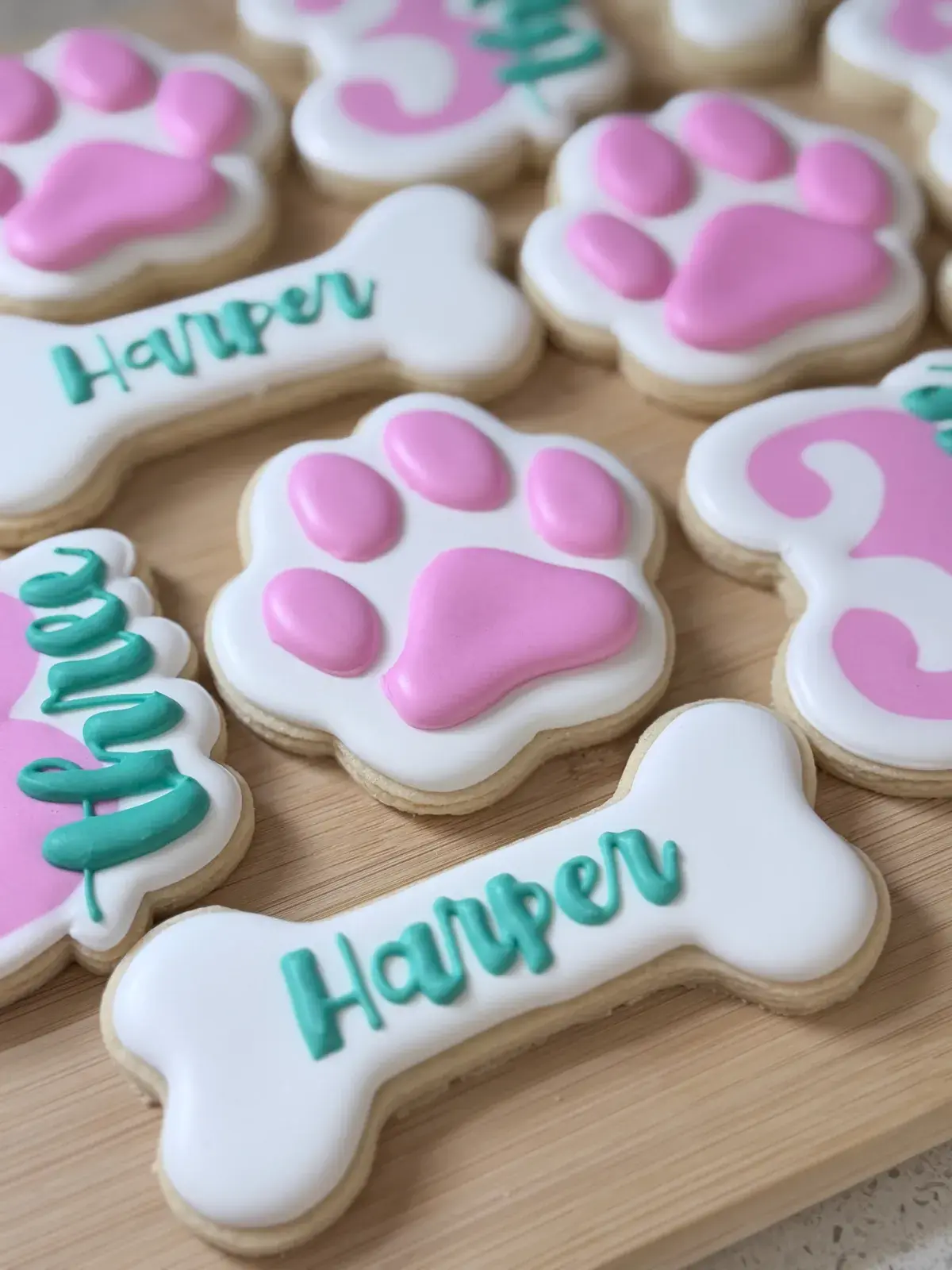 A closer view of the same puppy-themed cookie set, showing two bone-shaped cookies iced with the name Harper in teal script surrounded by pink-and-white paw print cookies.