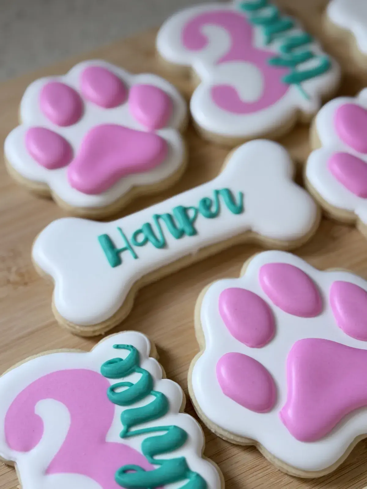 Close-up of a bone-shaped cookie iced with the name Harper in teal cursive script, flanked by pink-and-white paw print cookies and the edge of a pink number 3 cookie.