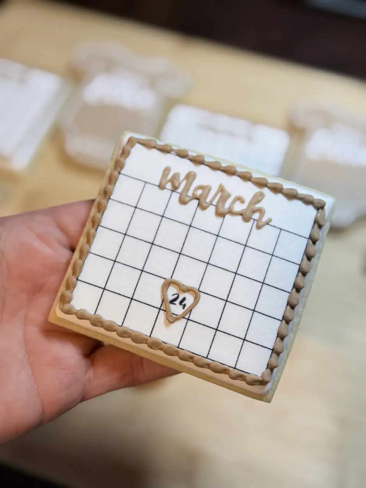 A hand holding a rectangular cookie iced to look like a March calendar page, with the date 24 circled in a piped heart.